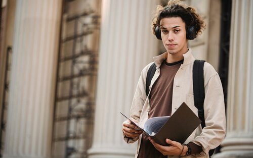 Nahaufnahme eines jungen Mannes mit dunklem Haar mit einer Akte in der Hand und Kopfhörern. | © pexels