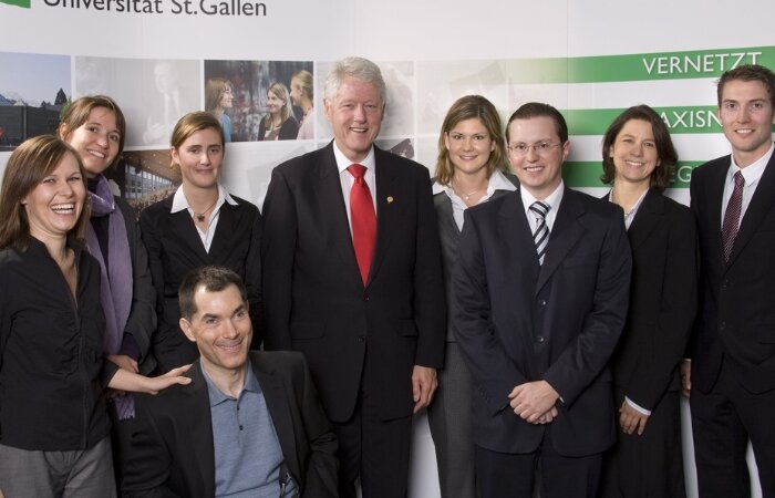 Eine Gruppe von Männern und Frauen, in der Mitte Bill Clinton mit roter Krawatte. Im Hintergrund das Logo der Universität St.Gallen.