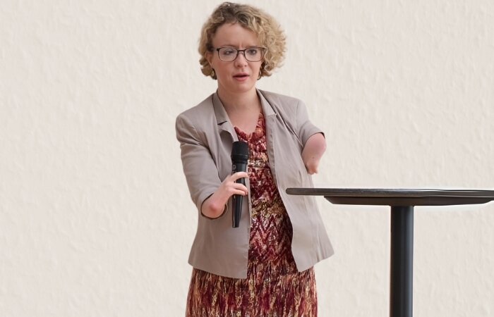 Foto von Debbie Stettler mit Mikrofon in gemustertem Kleid und Blazer, sie trägt eine Brille und hat blonde Locken.