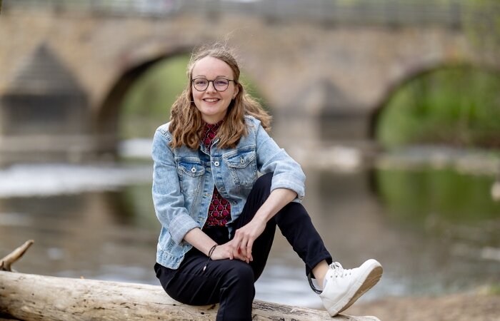 Elisa Bauch sitzt auf einer Mauer, hinter ihr ein Fluss und eine Brücke. Sie hat lange, helle Haare, trägt eine Brille und lächelt.