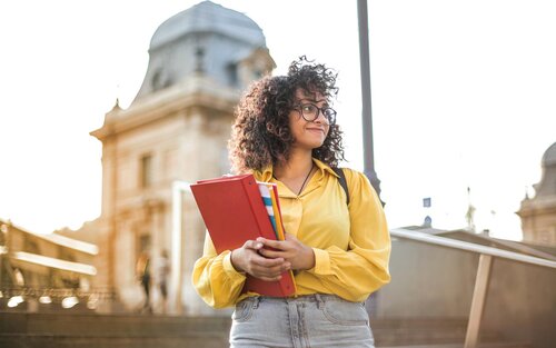 Une étudiante aux cheveux noirs frisés et portant des lunettes se tient devant une université, avec des livres dans les mains. Elle regarde au loin, un peu souriante, mais aussi un peu incertaine. | © pexels