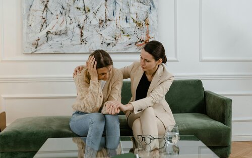 Zwei Frauen sitzen nebeneinander auf einem Sofa, die eine hat verzweifelt den Kopf in die Hände gestützt, die andere hält ihre Hand. | © pexels