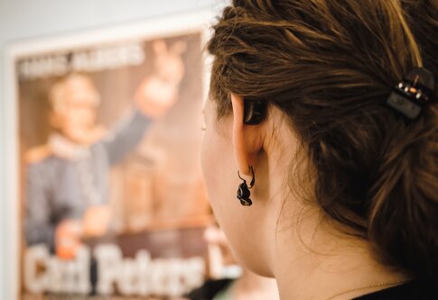 Une jeune femme avec un appareil auditif regarde une affiche floue. | © Gesellschaftsbilder.de
