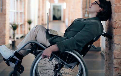 Foto einer Frau im Rollstuhl, die einen Laptop auf dem Schoss hat und resigniert an der Wand lehnt. | © pexels