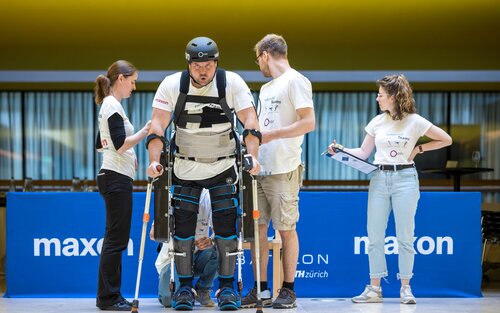 Ein Mann mit Helm, Exoskelett und Krücken in einer Arena. Neben ihm stehen zwei Frauen und Männern mit Klemmbrettern. | © ETH/CYBATHLON/Alessandro Della Bella