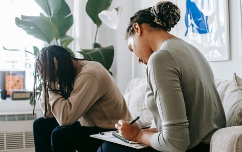 Ein Mann und eine Frau sitzen auf einem Sofa. Der Mann hat den Kopf in die Hände gestützt und sieht traurig aus. Die Frau hat einen Notizblock auf den Knien und macht sich Notizen. | © pexels